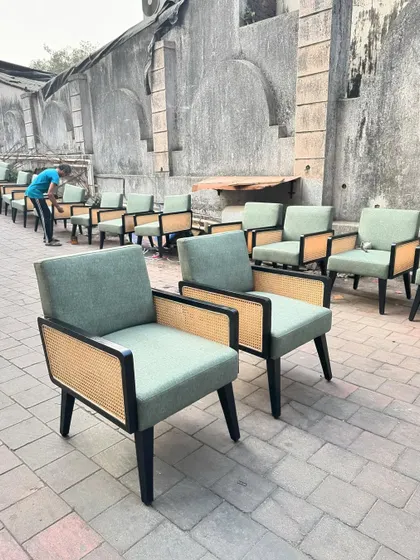 A large order of custom-made lounge chairs being prepared for a hospitality project. These feature a black-finished teak frame, natural cane side panels, and sage green upholstery.
