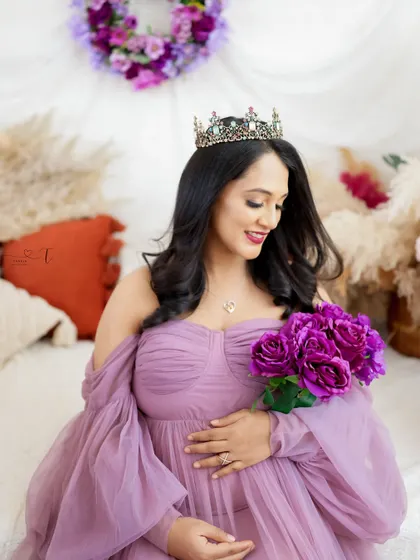 A beautiful portrait of a queenly mom-to-be. Wearing a tiara and a lavender gown, she holds a bouquet of purple flowers, looking serene and happy.