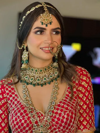 A picture of pure bridal elegance. Her makeup features a strong yet soft smokey eye, which contrasts beautifully with her traditional red and gold outfit and green kundan jewelry.