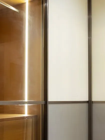 A detail shot of a sliding wardrobe where you can see the warm wood interior with LED lighting next to the frosted and grey glass panels. This contrast between the cool exterior and warm interior is a signature of my designs.