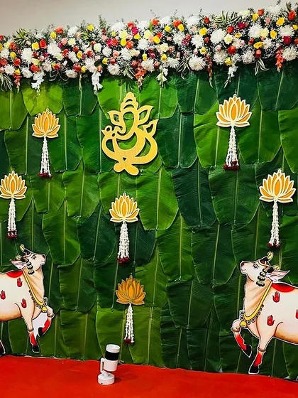 A traditional Seemantham backdrop featuring a wall of banana leaves, cow motifs, and a central Ganesha cutout. The decor is framed by colorful flower garlands for a complete festive look.