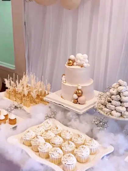 A dreamy dessert table with a two-tiered cake, cupcakes, and donuts, all in a soft white and gold palette. The fluffy cotton base makes it look like the treats are floating on a cloud.