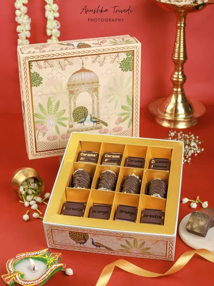 A beautifully designed peacock-themed chocolate box for Diwali, styled with diyas and jasmine flowers on a festive red backdrop.