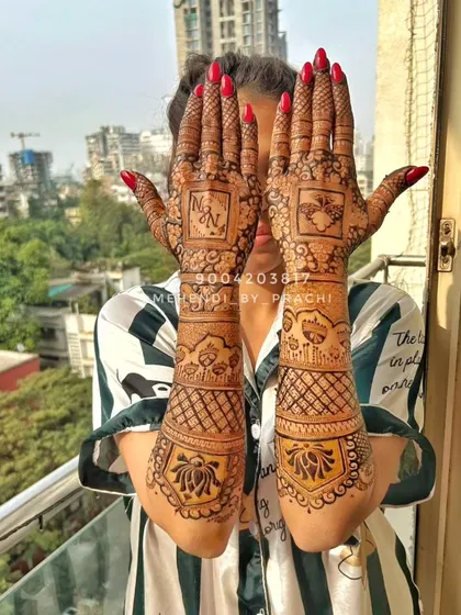 A personalized bridal mehndi with the couple's initials "N" and "N" prominently featured on the palms. The design is a mix of geometric checks and lotus motifs on the arms.
