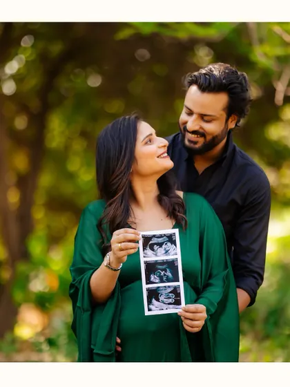 A happy couple shares their excitement in a beautiful park setting. The mother-to-be, dressed in a flowing green gown, proudly displays their baby's sonogram pictures for a lovely pregnancy announcement photo.