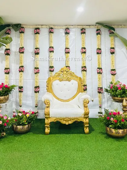 An elegant and minimalist stage setup. The clean white backdrop is adorned with hanging garlands of jasmine and pink roses, flanked by banana plants and brass pots filled with flowers.