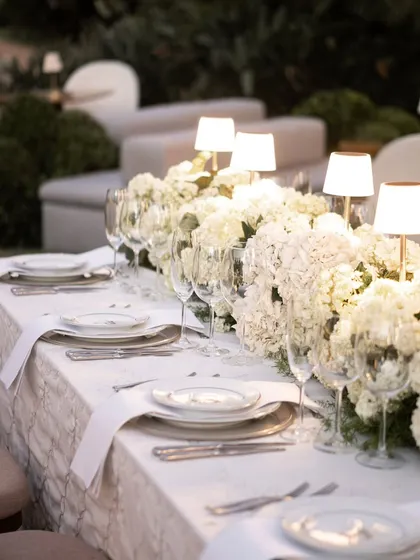 A close-up of a pristine white tablescape. The table is adorned with a thick runner of white hydrangeas and illuminated by the soft glow of individual table lamps, creating a luxurious and intimate dining setting.