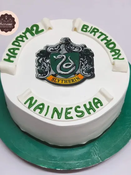 A Harry Potter Slytherin-themed cake. This simple design features the Slytherin house crest on a clean white whipped cream background, perfect for a dedicated fan.