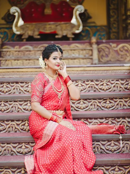 The intricate carving of the backdrop complements the traditional design of the red saree and gold jewelry.