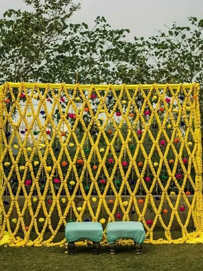 A beautiful photo backdrop made from a lattice of marigold garlands. The pops of colorful flowers woven into the yellow mesh add a playful and intricate detail to this outdoor setup.