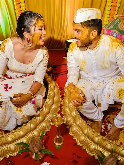 The bride and groom sharing a laugh while covered in turmeric and petals, seated in decorative urns during their joint haldi ceremony.