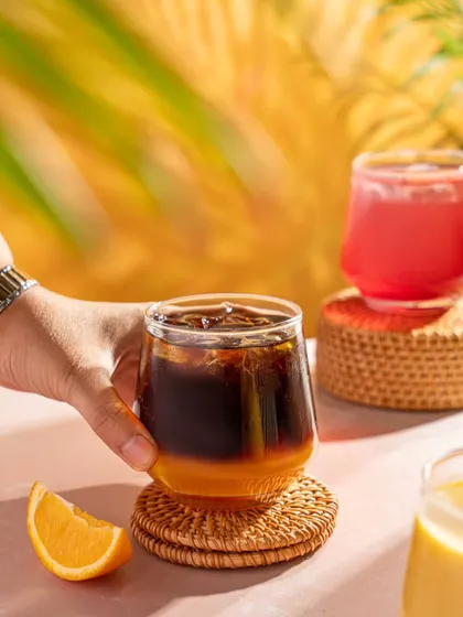 A hand holding a layered coffee and orange juice drink at Blue Tokai. The bright, tropical background and refreshing look of the drink make it perfect for a summer menu.