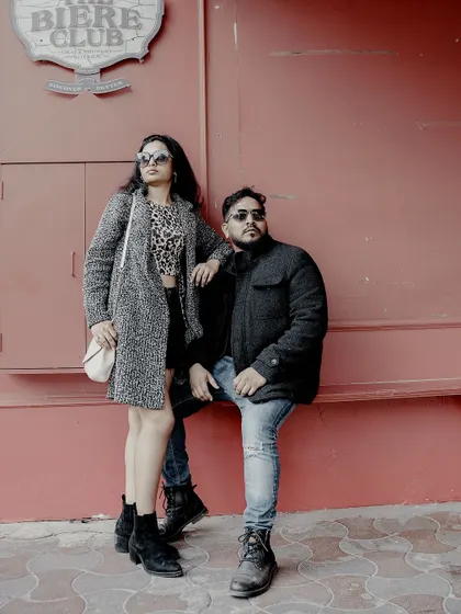 A full-length shot of a couple in stylish winter outfits, posing with attitude against a red wall.