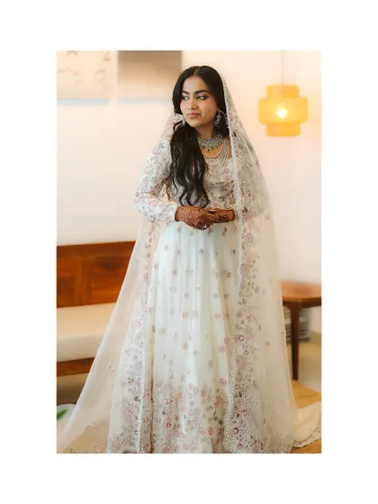 A bride in a magnificent white and pink embroidered gown stands in a softly lit room. Her regal posture and the intricate details of her dress create a portrait of pure elegance.