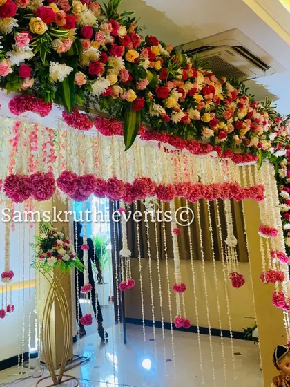 A beautiful floral entryway decoration. The arch is covered in pink and white flowers, with hanging garlands of jasmine and carnations creating a grand welcome for guests.