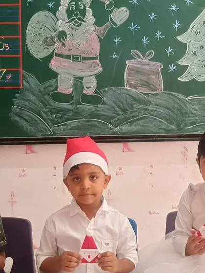 More students proudly display their Santa crafts in front of a Christmas-themed blackboard drawing.