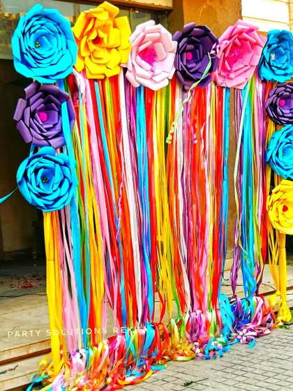A vibrant and colorful photo backdrop made of paper flowers and hanging ribbons. This is a fun and budget-friendly way to create a high-impact photo wall for a Mehendi or birthday party.