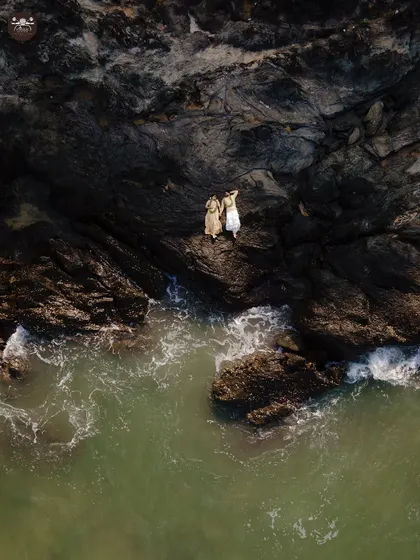 An aerial drone shot captures a couple standing on the edge of dramatic coastal rocks, with green waves washing ashore. This pre-wedding photograph highlights my ability to blend epic landscapes with intimate moments for a truly cinematic feel.