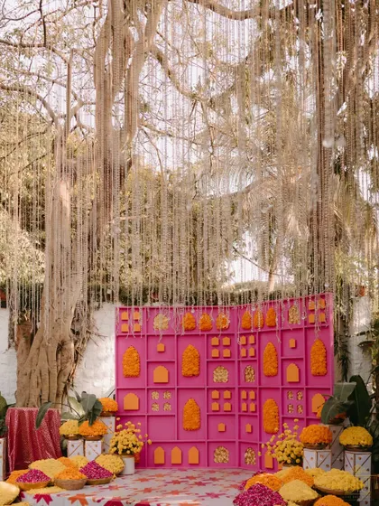 A bright pink backdrop decorated with marigolds and traditional motifs served as a perfect photo op at the Mehendi. Above, strings of pearls cascaded from a large tree, adding a touch of elegance to the vibrant setup.
