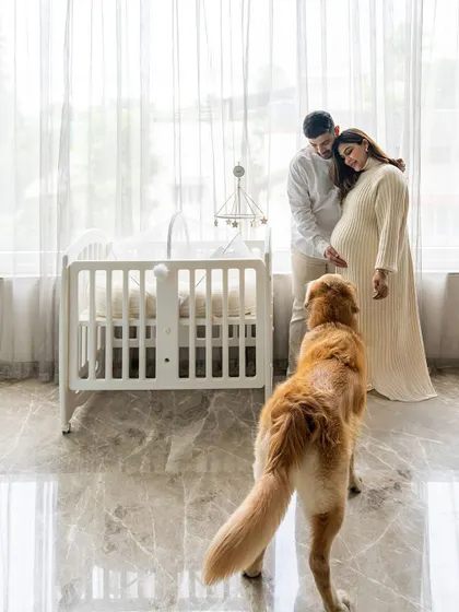 A full-length shot of an expecting couple by a crib, with their golden retriever looking towards them, creating a serene family scene.