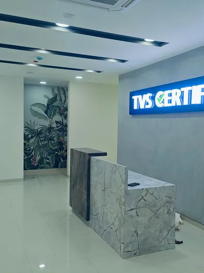 The reception desk at the TVS Certified center is crafted from contrasting marble-finish laminates. The backlit "TVS Certified" sign and concrete-texture wall create a modern and trustworthy brand presence.