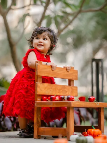 A baby girl in a vibrant red dress stands by a small wooden bench during her outdoor first birthday photoshoot.