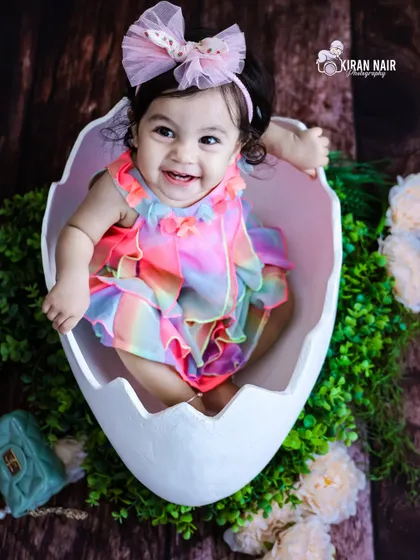 A joyful baby girl in a rainbow-colored outfit pops out of a large eggshell prop. This creative and colorful studio setup is perfect for a fun and unique baby photoshoot.