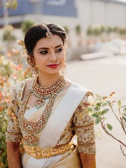 A beautiful outdoor shot of a bride in her traditional white and gold saree. The makeup features a classic red lip and defined eyes, a timeless choice for any bride.