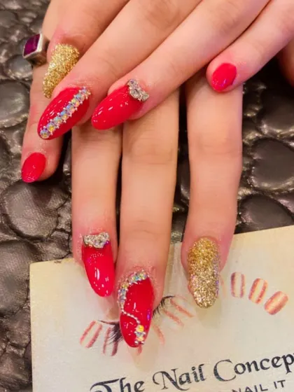 An extravagant set of red and gold almond nails. The design is packed with detail, including gold glitter, multi-colored rhinestones, and 3D bow embellishments.