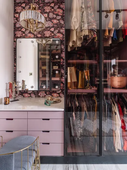 A full shot of the chic dressing area, complete with lilac-colored drawers, a vanity mirror with lights, and a glass-door wardrobe. The floral wallpaper adds a touch of romanticism.