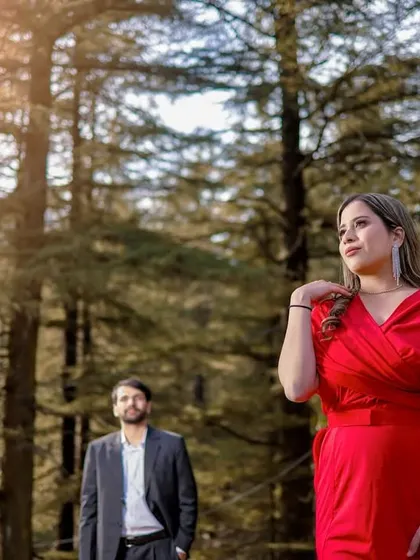 A beautifully composed shot of a woman in a red dress looking elegant in a sun-dappled forest, with her partner in the background.