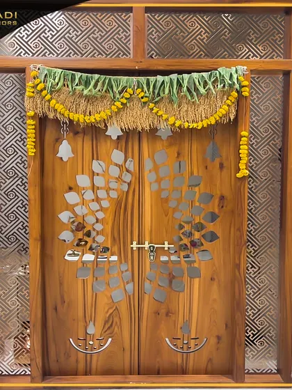 A beautiful wooden puja room door with a unique leaf-patterned metal inlay. The door is adorned with a traditional mango leaf and marigold toran, signifying auspiciousness.