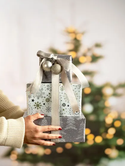 A lifestyle shot of hands holding a stack of beautifully wrapped Christmas presents. The blurred Christmas tree in the background creates a festive and heartwarming mood, perfect for a holiday campaign.