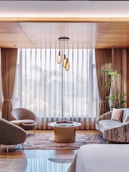 The sitting area in this hotel suite is defined by elegant, art deco-inspired metal screens that separate it from the sleeping area. This creates a multi-functional space that feels both open and private, enhanced by warm wood ceilings and soft lighting.