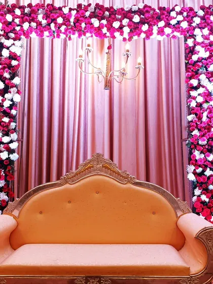 A close-up of a reception stage with a bright pink and white floral arch. The setup includes a vintage-style chandelier hanging in the center, set against a backdrop of pleated pink curtains for a rich, textured look.