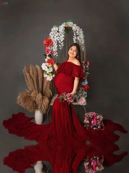A joyful portrait of a mom-to-be in a stunning red gown. She is seated on our floral swing prop, with the long train of the dress spread out beautifully around her.