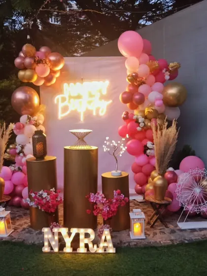A beautiful pink and gold themed birthday party. The decor includes a custom backdrop with a neon sign, metallic gold pedestals, a lush balloon garland, and personalized light-up letters.
