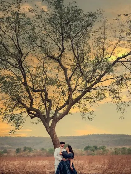 A stunning landscape shot featuring a couple under a large, dramatic tree against a sunset sky, creating a powerful and cinematic pre-wedding image.