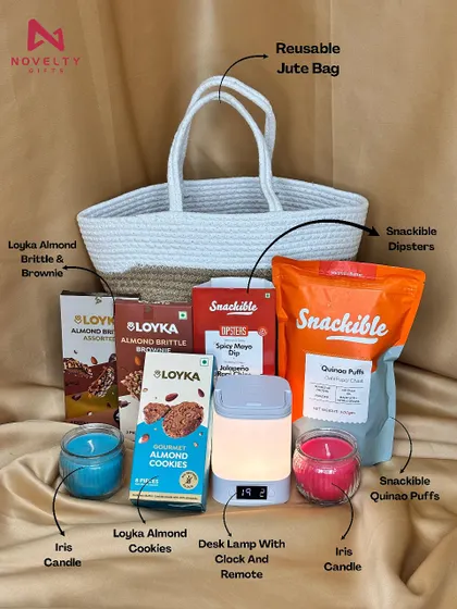 A closer look at a custom-designed hamper in a reusable jute bag. It's packed with treats like Loyka almond cookies, Snackible puffs, Iris candles, and a handy desk lamp with a clock.