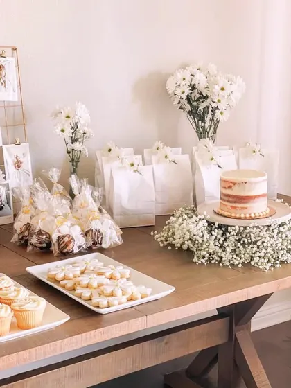 A rustic yet elegant dessert and favor table. The simple white cake, daisy-topped cookies, and fresh flowers create a charming and welcoming display for guests.