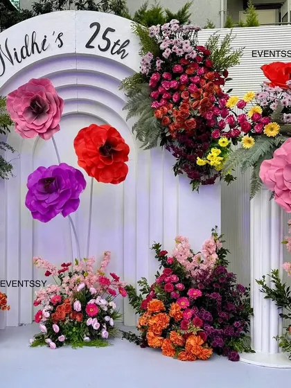 The central part of the 25th birthday backdrop, showing the custom "Nidhi's 25th" signage inside a modern arch. The arrangement of large paper flowers and fresh blooms is designed to look artistic and abundant.
