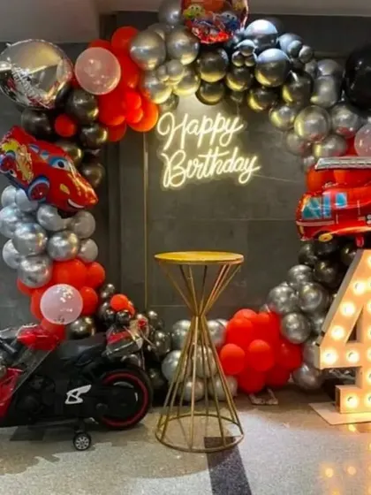 A 'Hot Wheels' and cars themed 4th birthday. The decoration includes a black, red, and silver balloon arch, car and fire truck foil balloons, a light-up number '4', and even a mini toy motorbike.