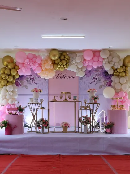 An elegant first birthday setup with a pink, white, and gold color scheme. The arrangement includes a large balloon arch, custom name backdrop, and a set of metallic-finish dessert tables and plinths.