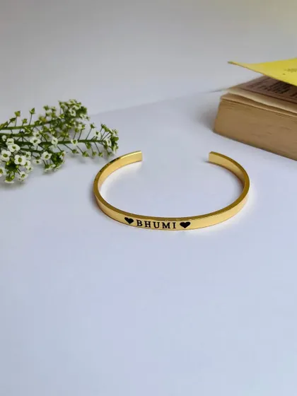 A single gold-plated Kada engraved with 'Bhumi' and hearts, displayed simply next to some flowers. This highlights the delicate and romantic feel of the piece.