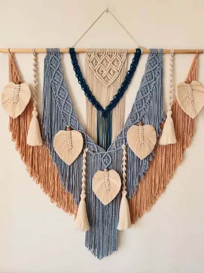 This custom macrame wall hanging was designed with a beautiful color palette of dusty blue, tan, and deep navy. The piece measures 40 inches wide and features multiple knotted patterns, tassels, and leaf details, making it a substantial and eye-catching work of art.