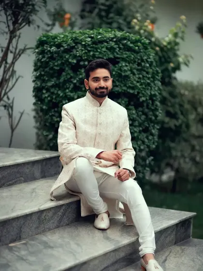 A stylish groom in a classic white sherwani, posing on the steps in a garden setting.