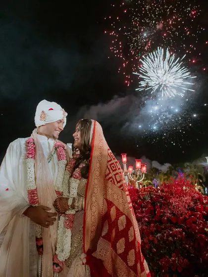 A nighttime shot of the couple with fireworks illuminating the sky. The decor, with its red flowers and glowing candelabras, creates a celebratory atmosphere that extends beyond the stage.