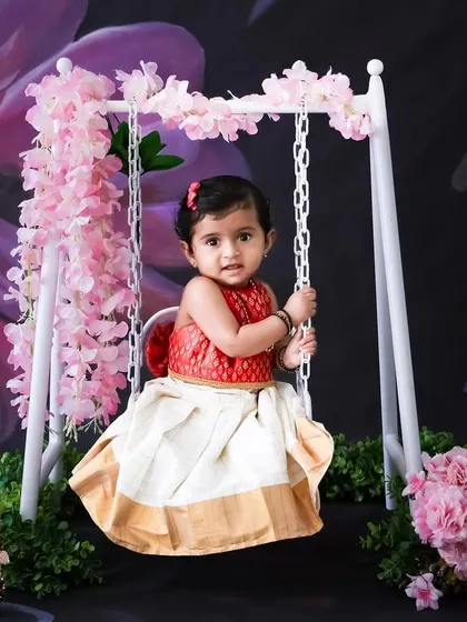 A toddler girl dressed in a traditional Onam pattu pavadai sits on a swing decorated with pink flowers. This shows my capability for creating beautiful setups for festive occasions.