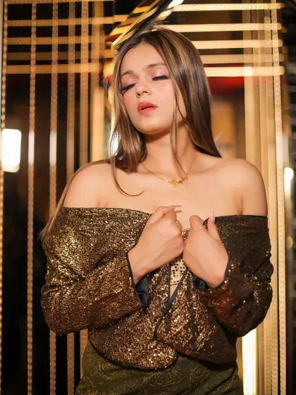 A glamorous studio shot of a model in a gold sequin off-shoulder top. The warm lighting and gold-toned background create a luxurious feel.