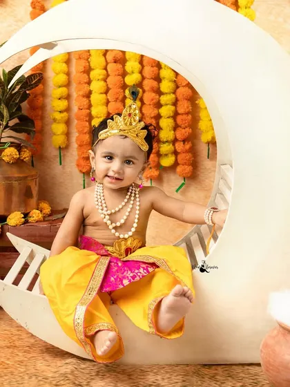 A sweet and smiling baby dressed as Krishna, posing perfectly on the moon prop.